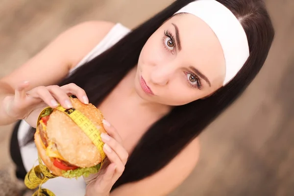 Une jeune femme tenant un hamburger dans un ruban à mesurer. Une fille se tient sur un fond en bois. La vue du haut. Le concept de saine alimentation . — Photo