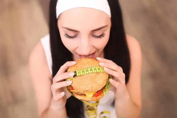 Une jeune femme tenant un hamburger dans un ruban à mesurer. Une fille se tient sur un fond en bois. La vue du haut. Le concept de saine alimentation . — Photo