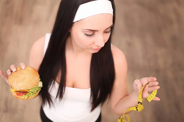 Eine junge Frau hält einen Burger und ein Maßband in der Hand. steht ein Mädchen auf einem hölzernen Hintergrund. die Aussicht von oben. das Konzept der gesunden Ernährung. — Stockfoto