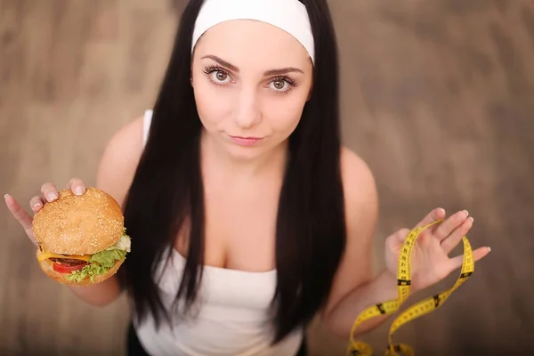 Eine junge Frau hält einen Burger und ein Maßband in der Hand. steht ein Mädchen auf einem hölzernen Hintergrund. die Aussicht von oben. das Konzept der gesunden Ernährung. — Stockfoto