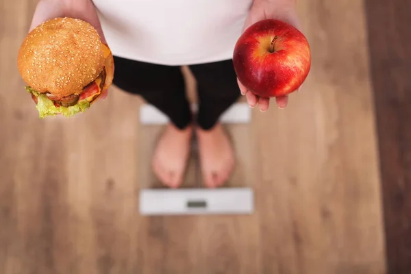 다이어트입니다. 여 자가 측정 몸 무게에 무게 규모 들고 햄버거 고 애플. 과자는 건강에 해로운 정크 푸드. 다이어트, 건강 한 식 생활, 라이프 스타일입니다. 체중 감량입니다. 비만입니다. 상위 뷰 — 스톡 사진