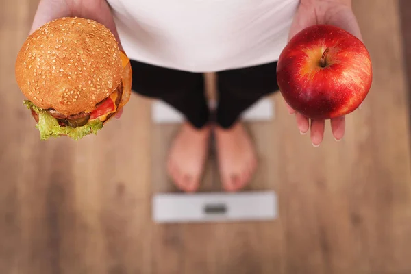 Kost. Kvinna mäta kropp vikt på väger skala Holding Burger och apple. Godis är ohälsosamma skräpmat. Bantning, sunda matvanor, livsstil. Viktminskning. Fetma. Ovanifrån — Stockfoto