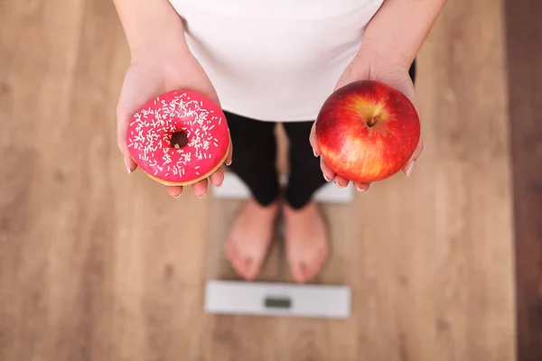 Δίαιτα. Γυναίκα μέτρησης σώματος βάρος για ζύγιση κλίμακας κρατώντας ντόνατ και μήλο. Τα γλυκά είναι ανθυγιεινά τρόφιμα παλιοπραγμάτων. Να κάνει δίαιτα, υγιεινή διατροφή, τον τρόπο ζωής. Απώλεια βάρους. Η παχυσαρκία. Το Top View — Φωτογραφία Αρχείου