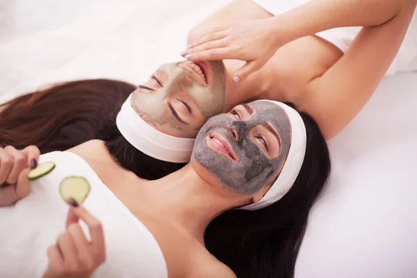 Spa en casa. Dos hermosas mujeres jóvenes sosteniendo pedazos de pepino en sus ojos y sonriendo mientras están en la cama — Foto de Stock