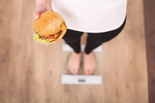 Concepto de dieta y comida rápida. Mujer con sobrepeso de pie en la escala de pesaje Holding Burger (Hamburger). Comida basura poco saludable. Dieta, estilo de vida. La pérdida de peso. Obesidad. Vista superior — Foto de Stock
