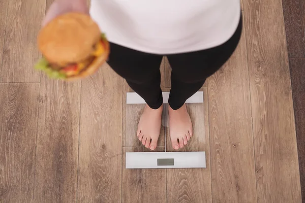 Conceito de dieta e fast food. Overweight Woman Standing On Weighing Scale Holding Burger (Hamburger) (em inglês). Unhealthy Junk Food. Dieta, Estilo de Vida. Perda de peso. Obesidade. Vista superior — Fotografia de Stock