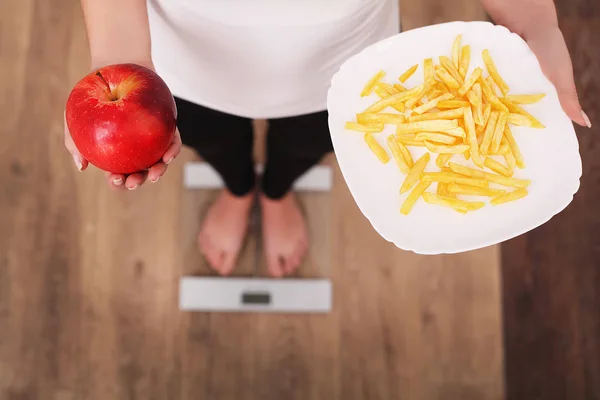 一个年轻漂亮的女人在磅秤上, 在苹果和薯片之间做出选择。健康饮食的概念。健康的生活方式。饮食. — 图库照片