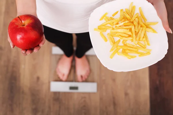 En ung vacker kvinna är på skalor och göra ett val mellan ett äpple och potatischips. Begreppet hälsosam kost. Hälsosam livsstil. Kost. — Stockfoto