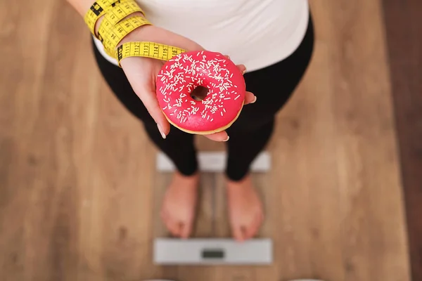 Young beautiful woman standing on scales and holding a donut. The concept of healthy eating. Healthy Lifestyle. Diet.