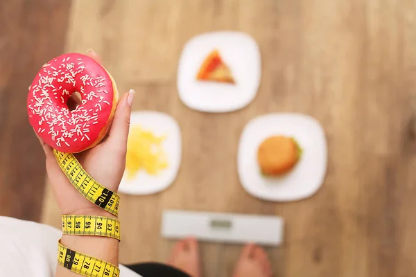 Ölçekler üzerinde duran ve donut tutan genç güzel kadın. Sağlıklı beslenme kavramı. Sağlıklı bir yaşam tarzı. Diyet. — Stok fotoğraf