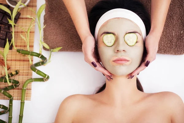 Beautiful young woman is getting facial clay mask at spa, lying — Stock Photo, Image