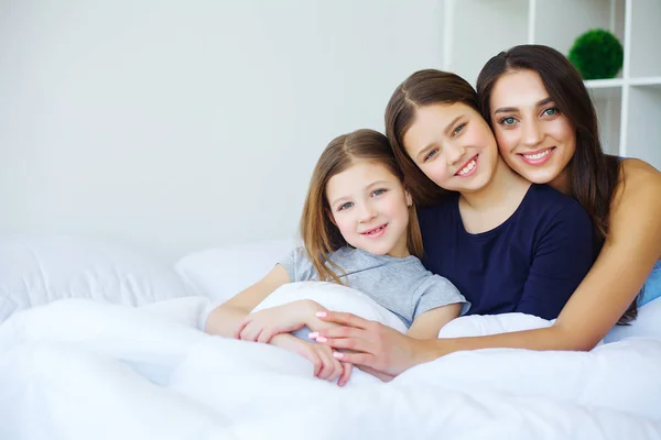 Beautiful mom and daughters are hugging, looking at camera and s