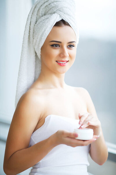 Young woman standing in bathroom and applying face cream in the 