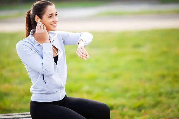 Foto de Joyful Fitness Woman 30s en ropa deportiva tocando el auricular Bluetooth y sosteniendo el teléfono móvil, mientras descansa en Green Park — Foto de Stock
