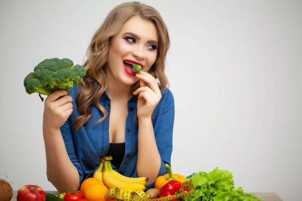 Cute woman at a table holding a broccoli on a background of fruit and vegetables — Stock Photo, Image