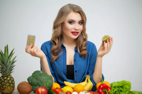 Kvinna vid ett bord som håller en kiwi och piller på en bakgrund av frukt och grönsaker. — Stockfoto