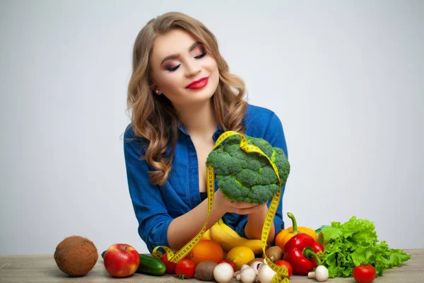 Kvinna vid bordet håller en broccoli på bakgrund av frukt och grönsaker — Stockfoto