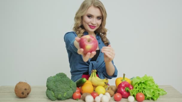 Nette Frau an einem Tisch mit einem Apfel auf einem Hintergrund aus Obst und Gemüse. — Stockvideo