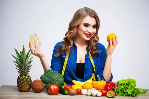 Mujer en una mesa sosteniendo un kiwi y pastillas sobre un fondo de frutas y verduras — Foto de Stock