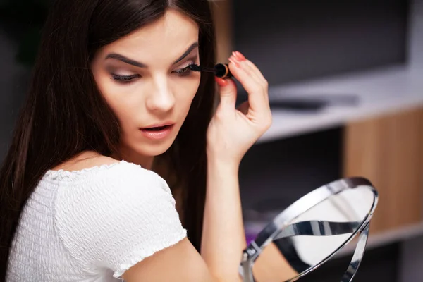 Chica bonita aplica maquillaje en la cara en casa cerca del espejo —  Fotos de Stock