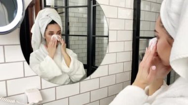 Genç bir kadın beyaz önlüklü banyoda makyaj malzemesi kullanıyor.