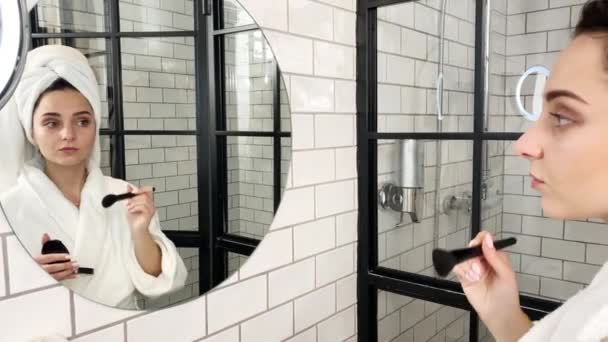 Mujer joven aplicando cosméticos en la cara en el baño con abrigo blanco — Vídeo de stock