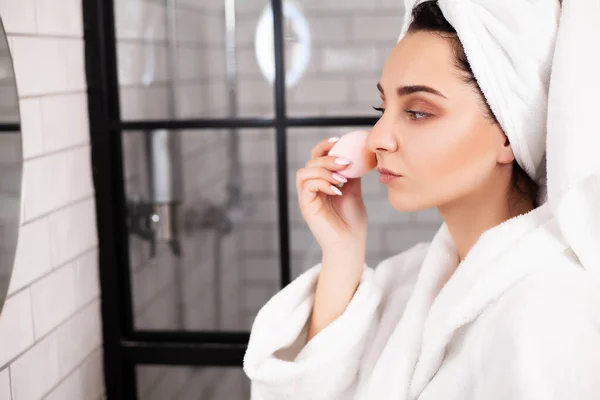 Vrouw in de badkamer met een handdoek op haar hoofd brengt make-up op haar gezicht — Stockfoto