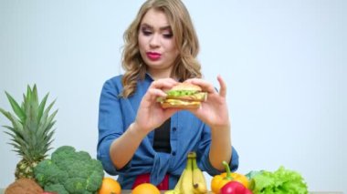 Güzel bir kadın mutfakta elinde kötü niyetli bir burgerle taze sebzeler ve meyvelerle bekliyor.