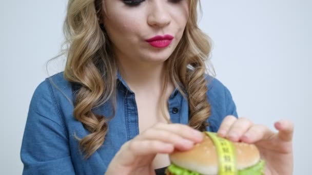 Mujer bonita en la cocina sosteniendo una hamburguesa maliciosa sobre un fondo de verduras y frutas frescas — Vídeo de stock