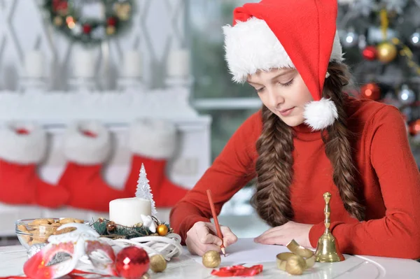 クリスマスのテーブルに座っての準備サンタ帽子で幸せな少女の肖像画 — ストック写真