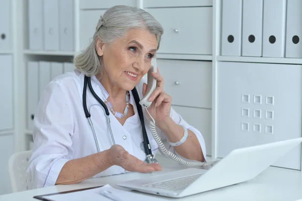 Senior Femme Médecin Parler Téléphone Tout Travaillant Table Avec Ordinateur — Photo