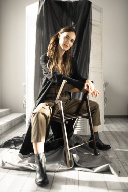 Moda tarzı giyinmiş çekici esmer kadın sandalyede oturuyor ve stüdyoda poz veriyor. 