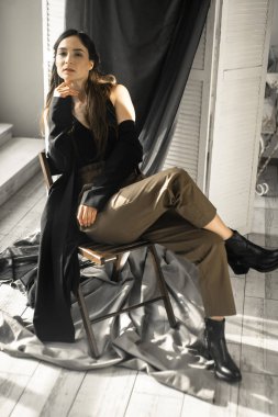 Moda tarzı giyinmiş çekici esmer kadın sandalyede oturuyor ve stüdyoda poz veriyor. 