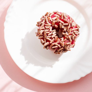 Çörek kırmızı sır ile kaplı ve gölgeler in numune yüzeyi ile doğal ışık altında pembe arka plan üzerinde beyaz tabakta rendelenmiş çikolata ile serpilir