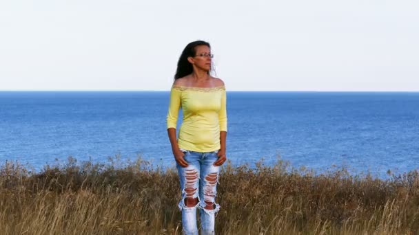 4K. Mujer adulta en blusa amarilla de pie en la orilla del mar. Día de verano — Vídeos de Stock