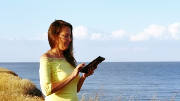 4k. Erwachsene Frau in gelber Bluse arbeitet an einem Sommertag am Meer mit Tablet — Stockvideo