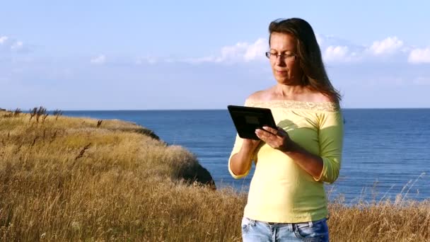 4K Las vacaciones modernas de la mujer madura armoniosa con la tableta contra el mar — Vídeo de stock