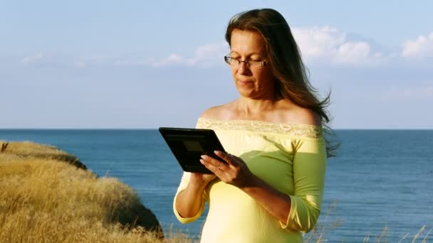 4K.Modern vacaciones de la mujer madura armoniosa con la tableta contra el mar — Vídeo de stock