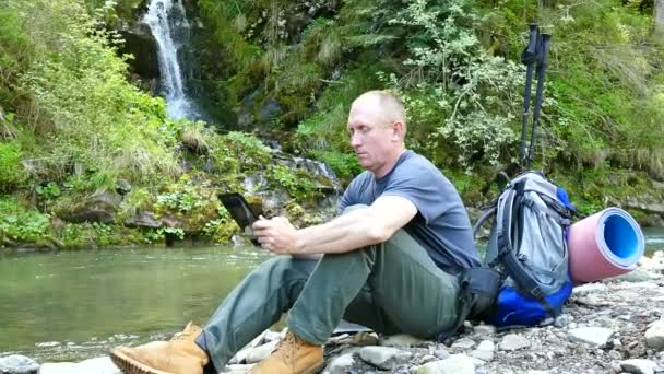 4K.. Hombre turista cerca de la computadora tableta de trabajo cerca del río y la cascada. Cambio de enfoque — Vídeo de stock