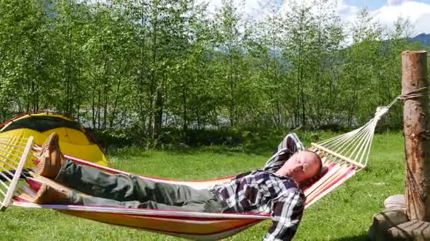 4K. Чоловік в жовтих дужках спить і трясеться в гамаку проти туристичного намету — стокове відео