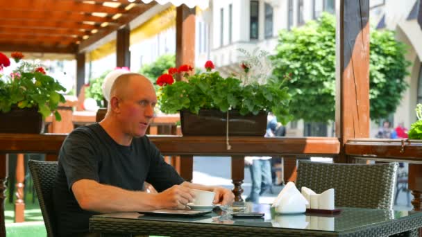 4К. Взрослый мужчина в уличном кафе с чашкой кофе разговаривает мобильный телефон — стоковое видео