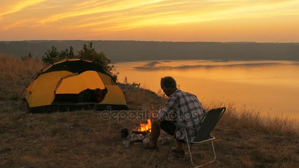 4K  Tent, bonfire, sunrise and man traveller near lake in sunrise time. — Stock Video