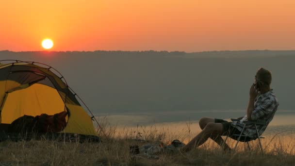 4K Tenda, fogueira, nascer do sol e homem viajante fala por telefone móvel — Vídeo de Stock