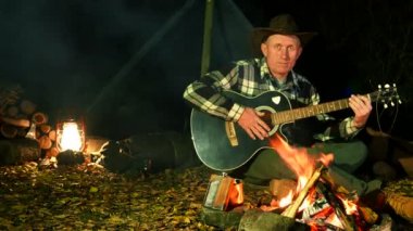  4k. Şenlik ateşi yakınındaki kovboy şapkalı adam gitar çalıyor. Sonbahar Seyahat hayat. 