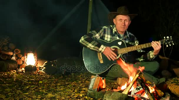 4K.Man en sombrero de vaquero cerca de hoguera toca la guitarra. Vida de viaje de otoño . — Vídeo de stock