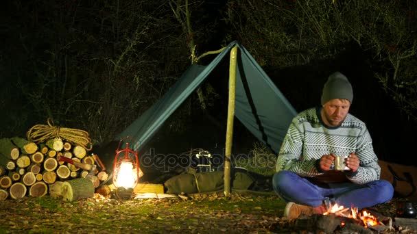 4k. Un jeune homme boit du café ou du thé au feu de camp. Vie en voyage, temps du soir — Video