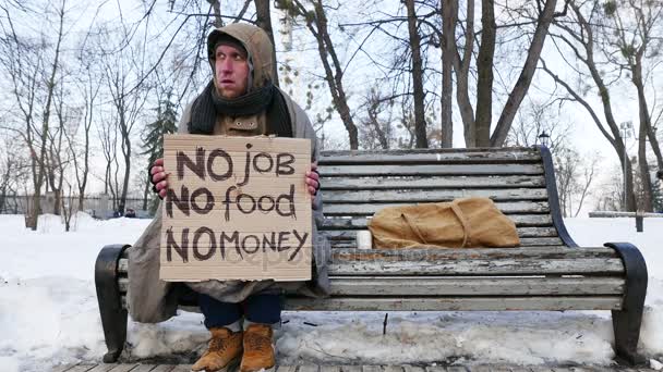 4K. Desesperación de los desempleados. Hombre con cartón sentarse en el banco en el parque de invierno — Vídeos de Stock