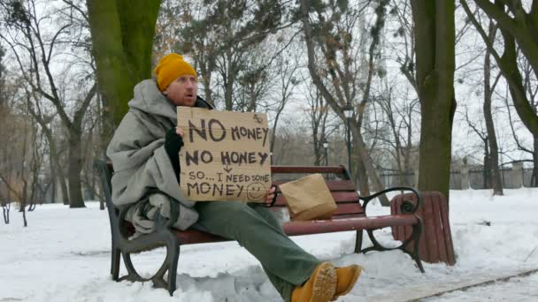 4K. Дорослий бездомний бідний чоловік з картоном, сидить на зимовій міській лавці — стокове відео