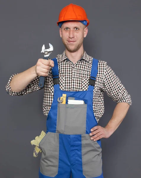 Sorrindo jovem trabalhador ou encanador em capacete segurar chave contra fundo cinza — Fotografia de Stock