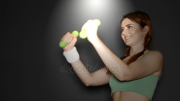 Atractiva mujer deportiva entrena con pesas delante de fondo oscuro — Vídeos de Stock